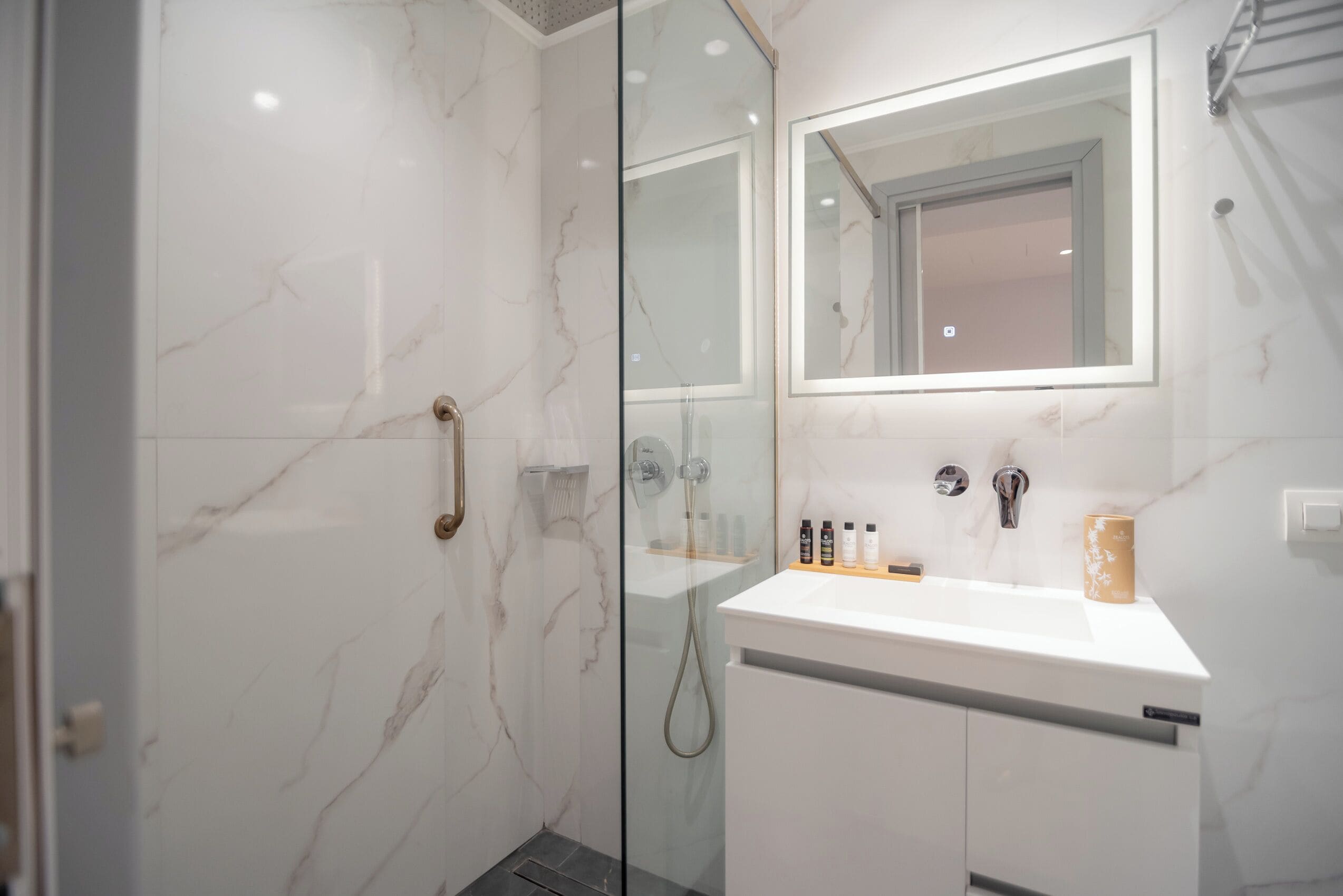 Un baño moderno con ducha de vidrio, un lavamanos blanco con espejo y artículos de aseo sobre el lavamanos.