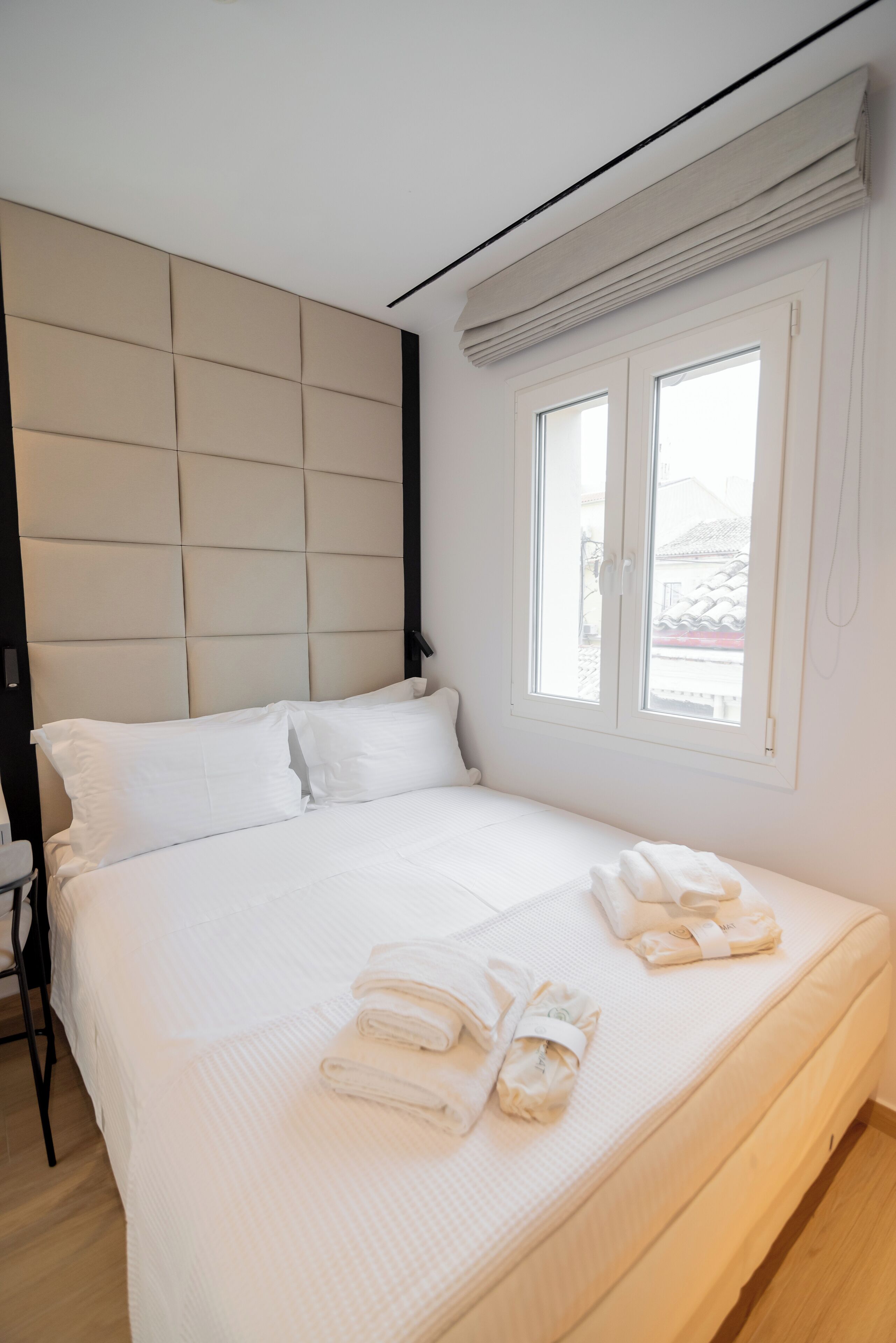 A neatly made bed with white linens and towels, a headboard, and a window with blinds.