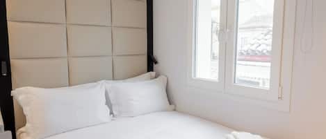 A neatly made bed with white linens and towels, a headboard, and a window with blinds.