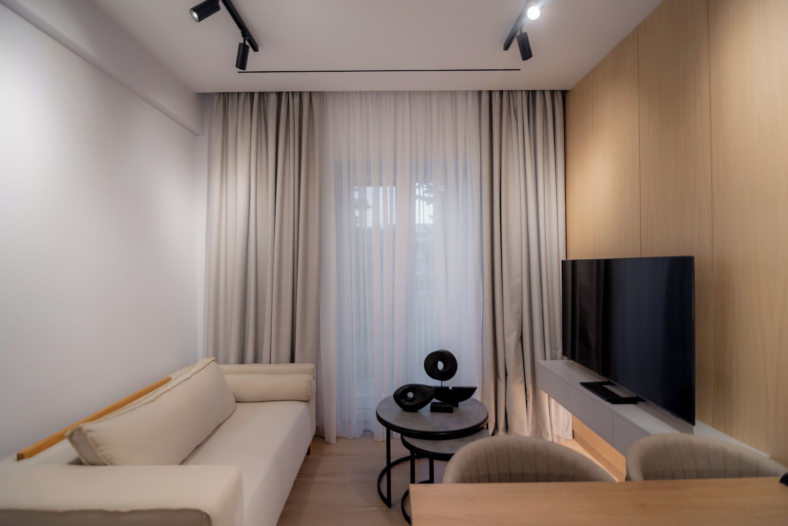 Ein modernes Hotelzimmer mit einer Couch, einem kleinen runden Tisch mit einem Lautsprecher, einem an der Wand montierten Fernseher und einem großen Fenster mit Vorhängen.