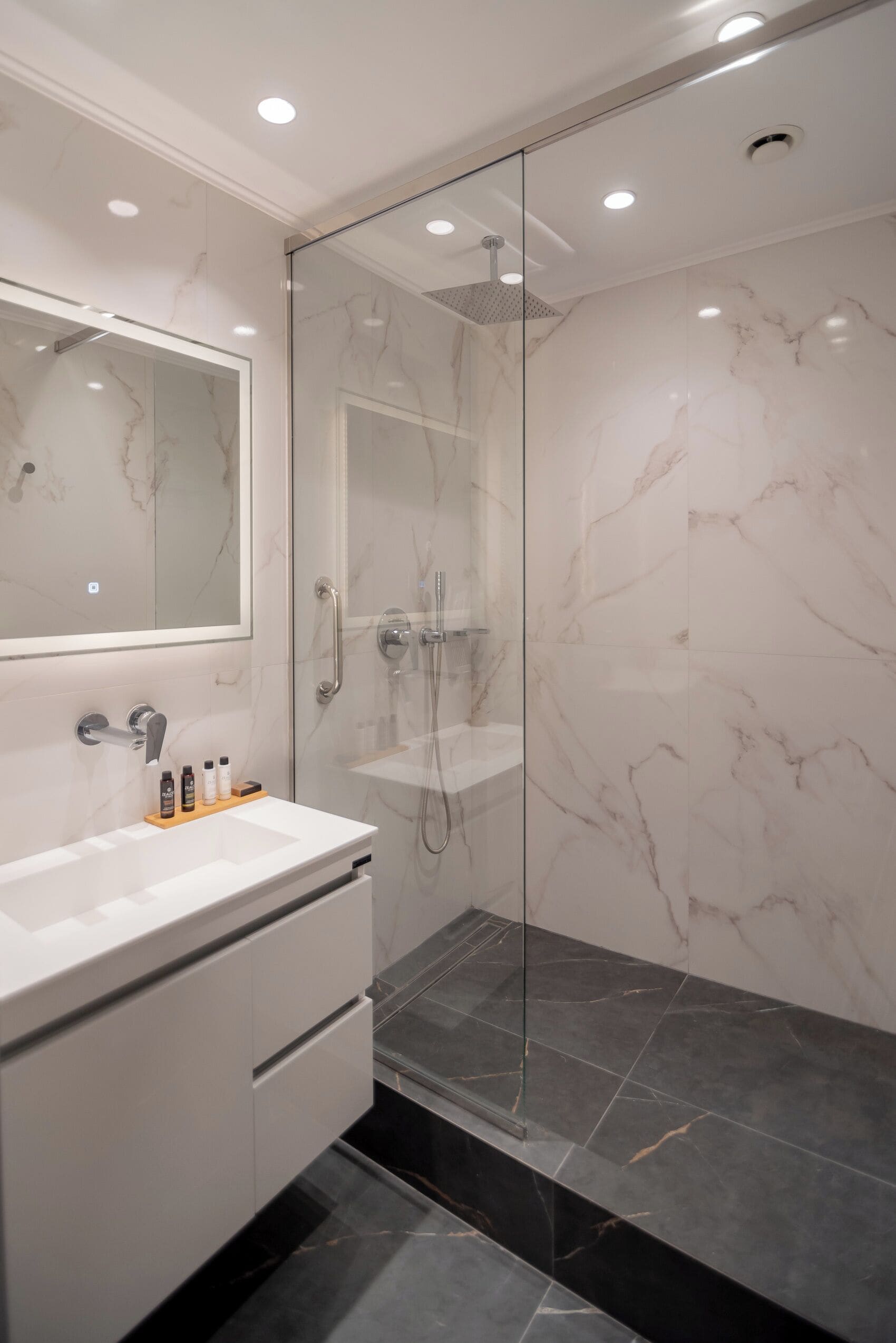A modern bathroom with a glass-enclosed shower, a marble-tiled wall, and a white vanity with a mirror and toiletries.