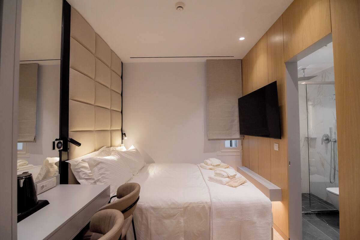 A modern hotel room with a large bed, a bedside table with a lamp, a chair, a flat-screen TV, and a glass-enclosed shower area.