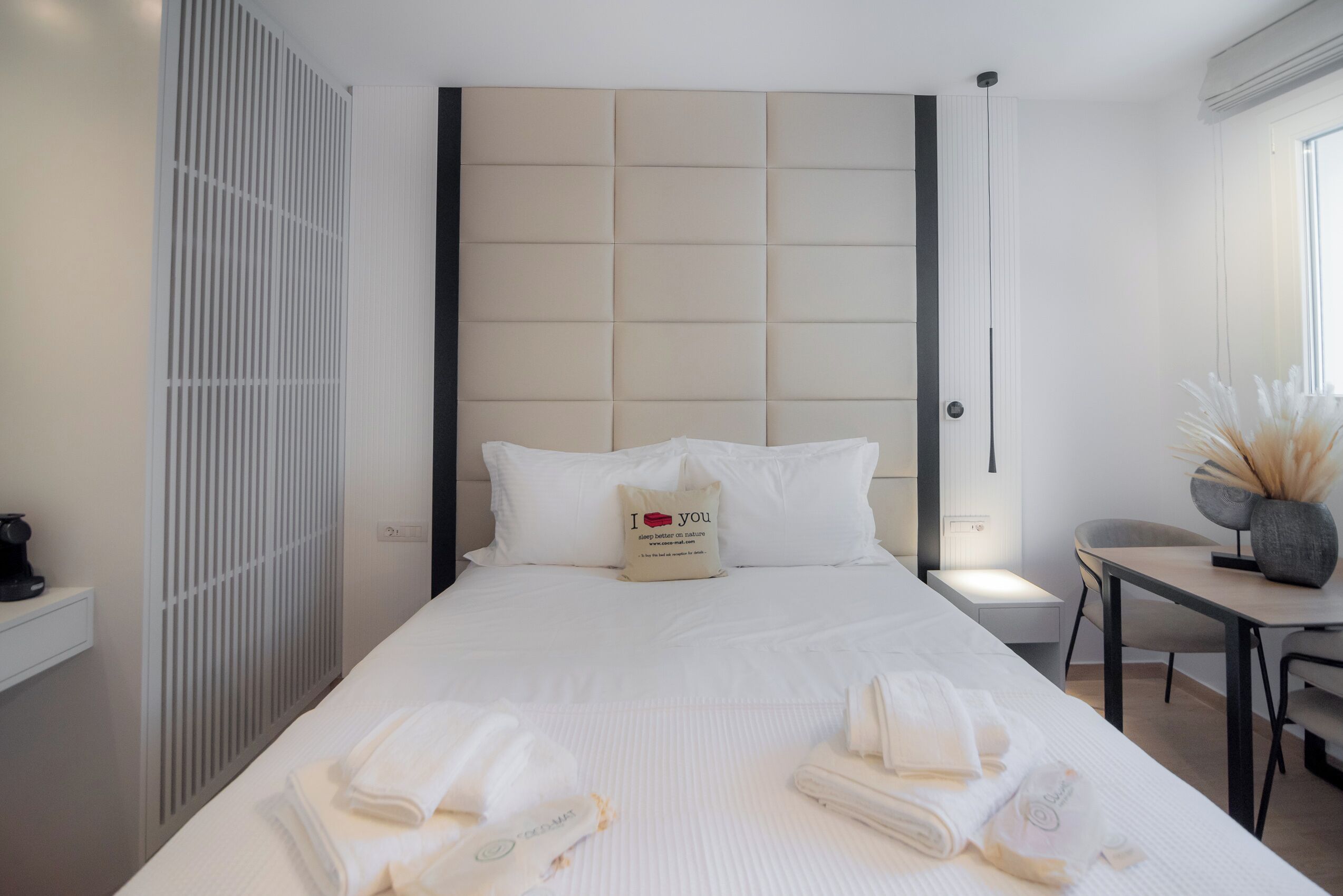 A modern hotel room with a large bed, a bedside table, a chair, and a small desk with a vase of dried flowers.