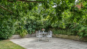 A patio with a table and chairs, surrounded by lush greenery and a brick wall.