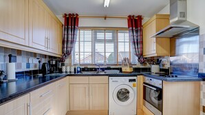 A compact kitchen with wooden cabinets, a built-in washing machine, a stove, and a window with curtains. - Swallowdale | East Ruston Cottages (Trunch)