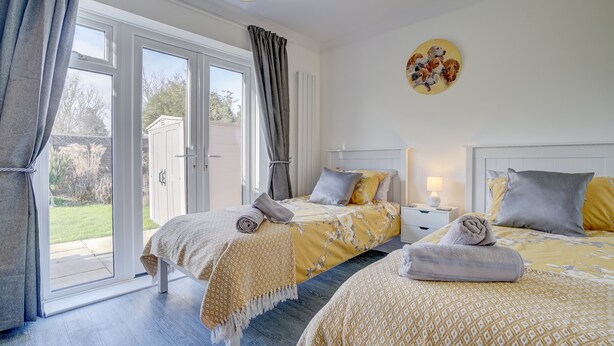 A bedroom with two beds, a sliding glass door, and a wall with a circular painting. - Swallowdale | East Ruston Cottages (Trunch)