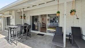 An outdoor patio area with a table and chairs, a sliding glass door leading to a covered entrance, and hanging plants.