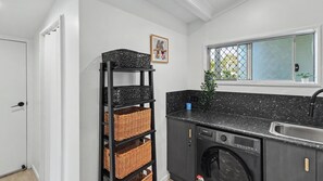 A compact laundry room with a washing machine, a tall wicker basket shelf, and a window with a view. - Vitamin Sea - Rainbow Beach (Rainbow Beach)