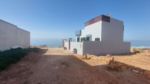 Un edificio moderno con vista sul mare, un sentiero sterrato e un muro di pietra.