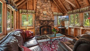 A cozy log cabin interior with a stone fireplace, leather furniture, and large windows offering views of the outdoors.