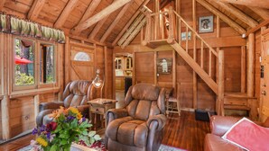 A cozy log cabin interior with a loft bedroom, leather armchairs, a dining area, and a staircase.
