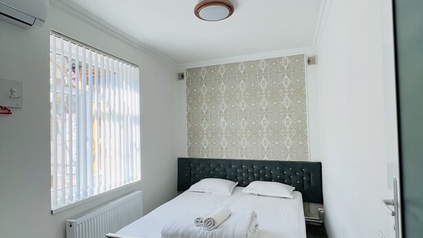 Une chambre à coucher avec un lit, un radiateur, une fenêtre avec des stores et un mur décoré d’un motif.