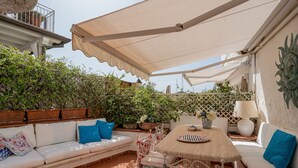 Een buitenterrein met een terras, een witte bank, een tafel en stoelen. Er staan potplanten en een decoratief wandobject.