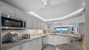A modern kitchen with white cabinetry, a granite countertop, a built-in microwave, a toaster, and a view of the ocean through large windows. - 🩱CBC 1-15E – Private balcony, beachfront, pool, gym & pickleball! (Clearwater)