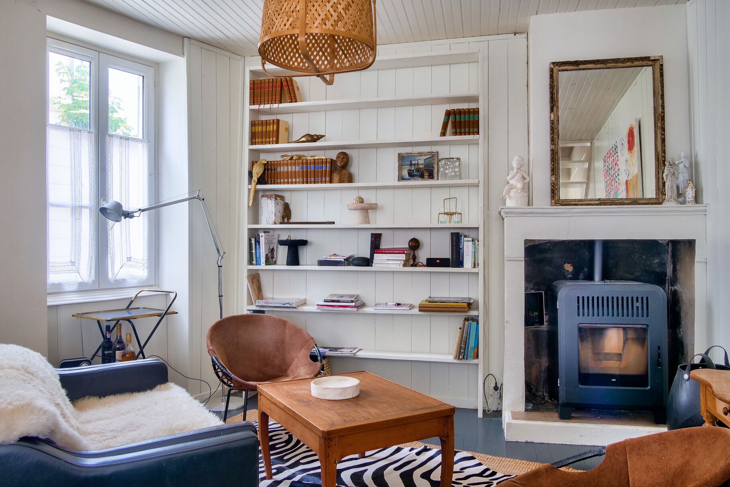 Un salon chaleureux avec une cheminée, des étagères remplies de livres et d’objets décoratifs, un fauteuil confortable et une table sur laquelle se trouve un bol.