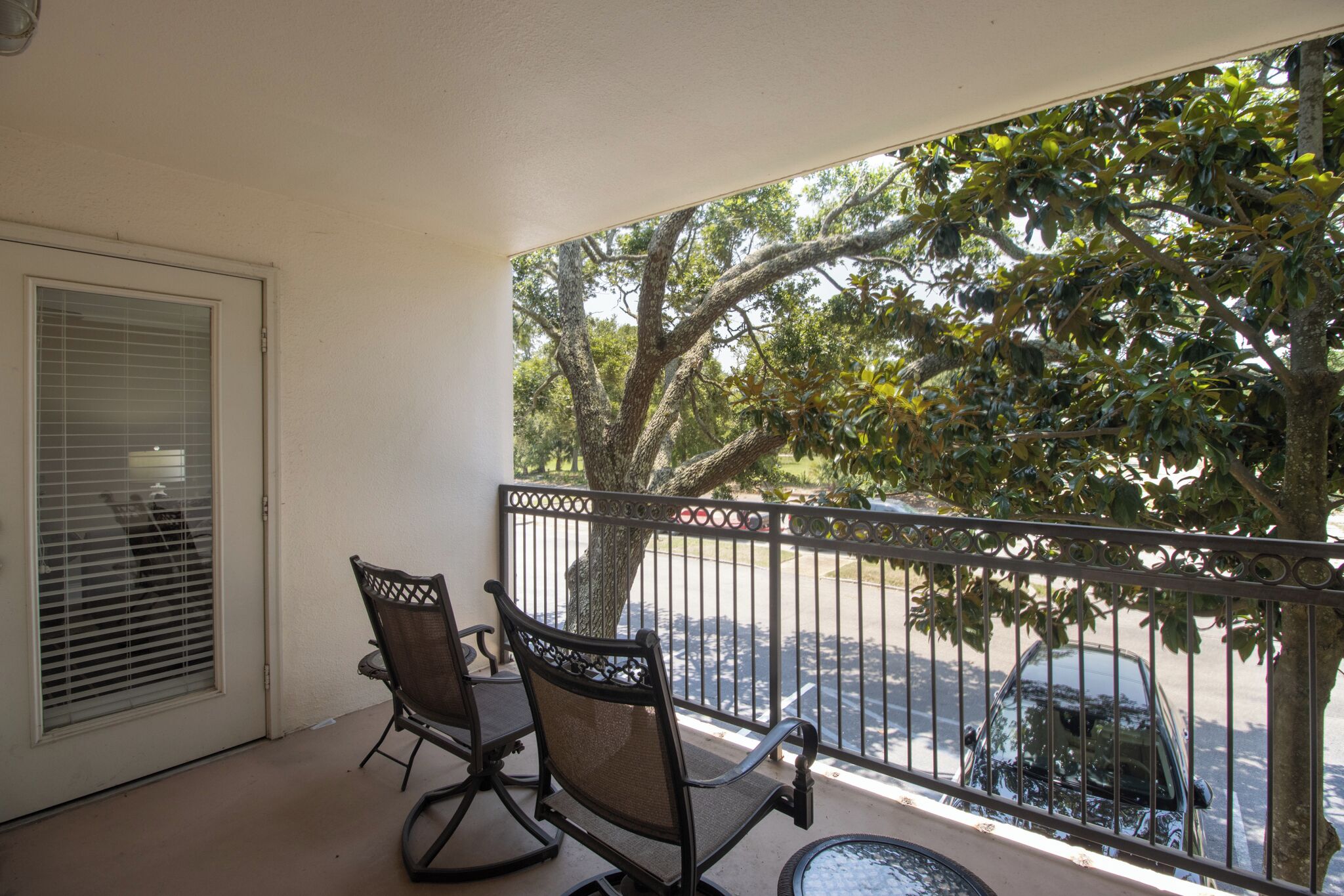 A balcony with two chairs and a table, overlooking a street with parked cars and trees.