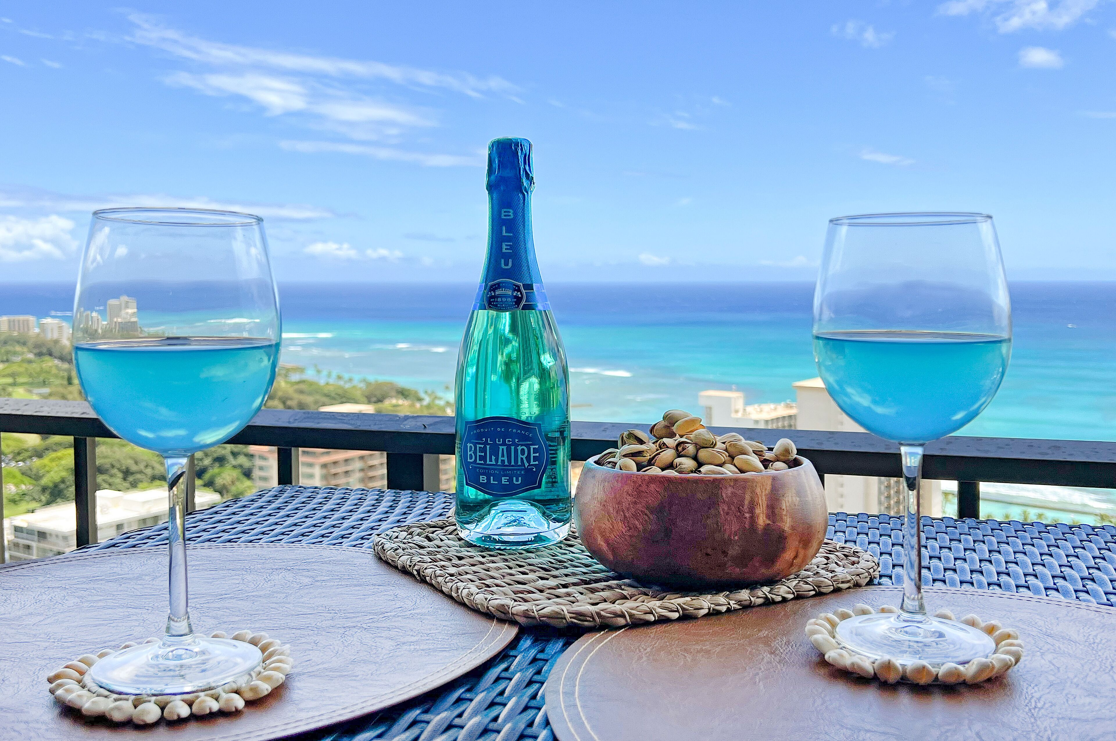 A table with two glasses of blue liquid, a bottle of Bel Air, and a bowl of nuts on a balcony overlooking a coastal view.