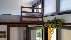 A bunk bed with a wooden frame, a white mattress, and a pillow. There is a window with blinds, a door, and a hanging plant. - Tiny Villa among the Olives. (hERAKLION)