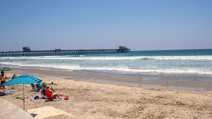 A beach with people relaxing under umbrellas, a pier extending into the ocean, and a sandy shore. - Seaside Serenity: Enjoy Ocean Views from Your Patio! (Oceanside)