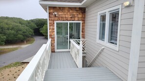 A wooden deck with a glass door leading to a covered outdoor area, a white railing, and a view of a road and trees.