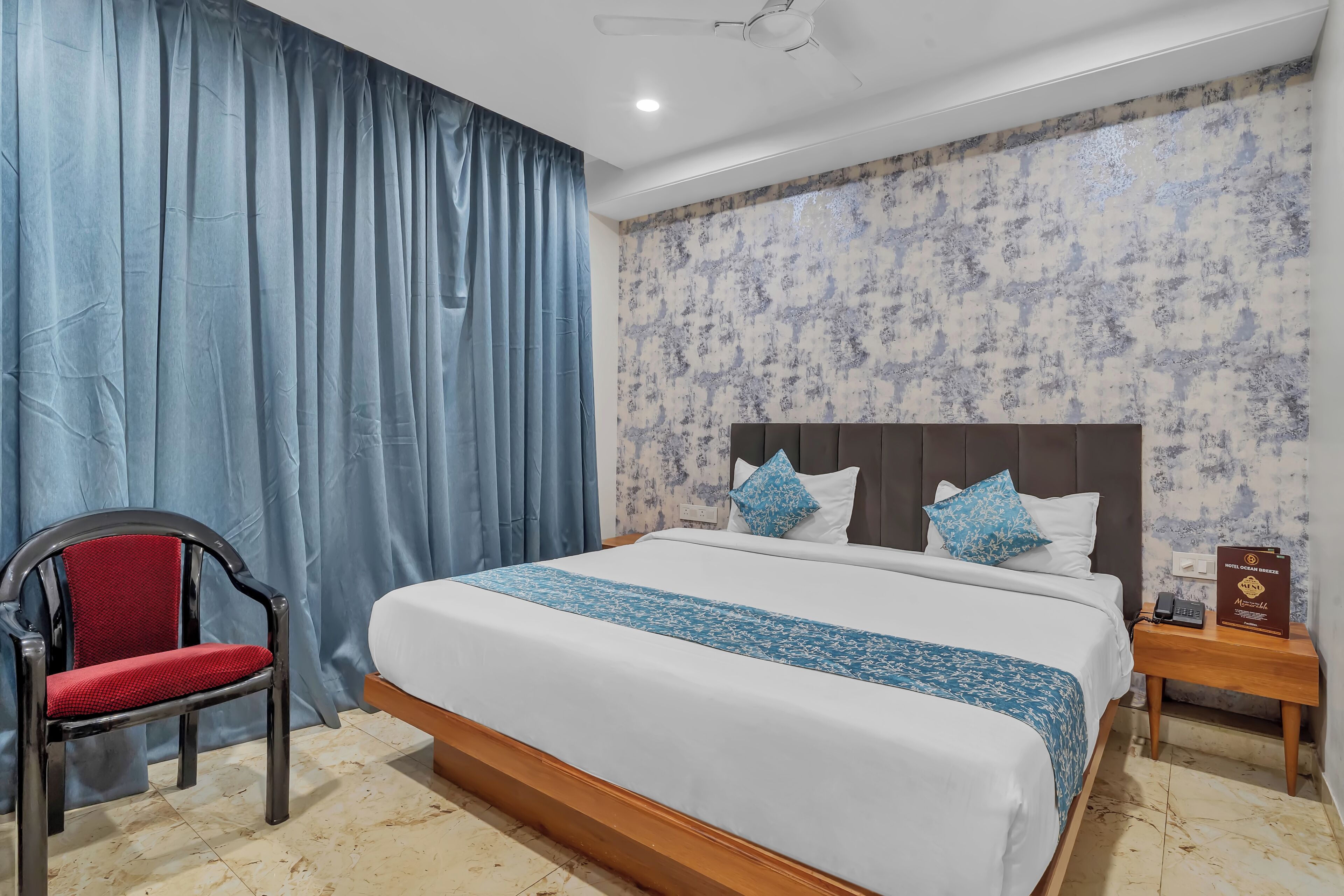 A hotel room with a large bed, a chair, a nightstand, and a wall with a floral pattern.