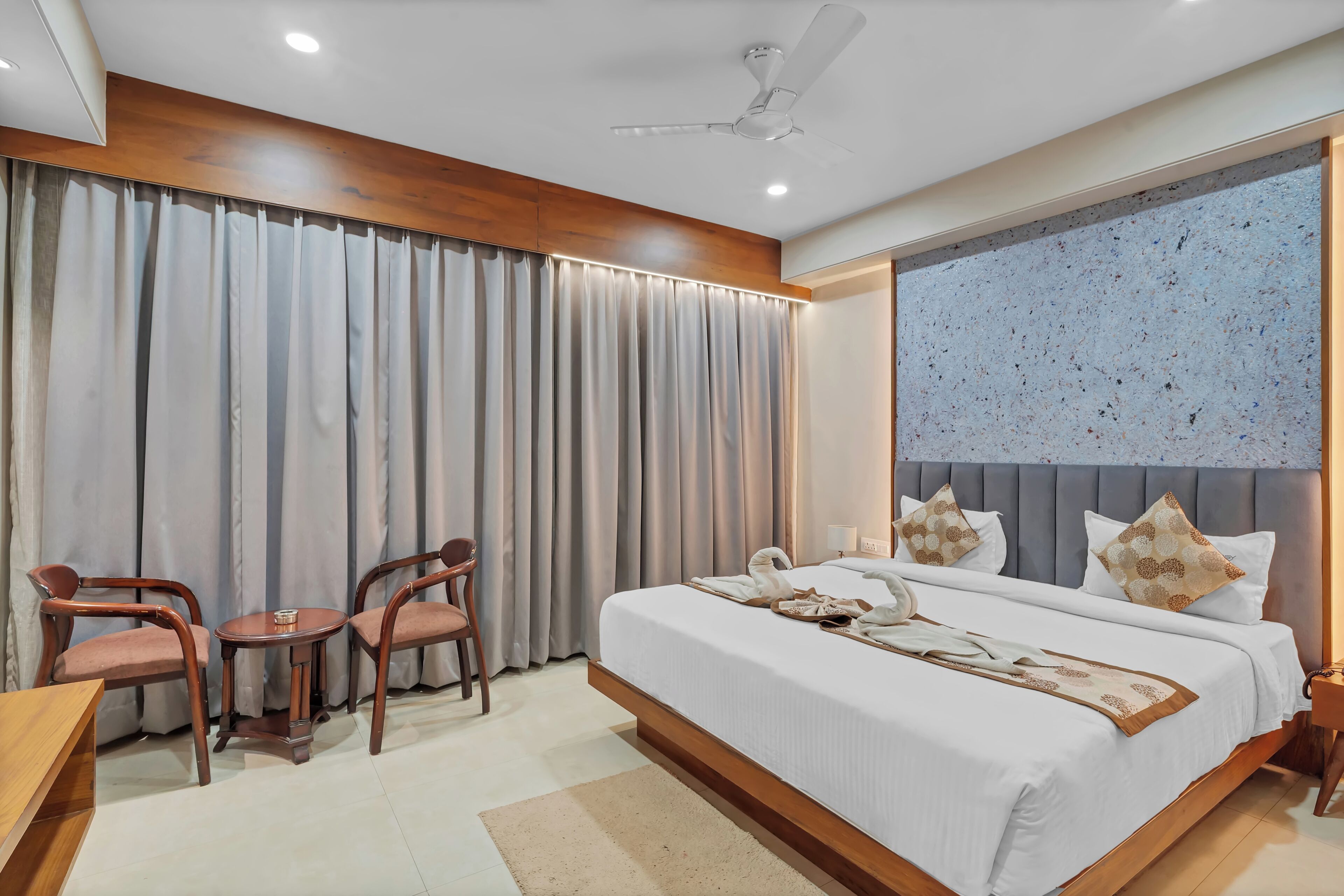 Quarto de hotel moderno com uma cama grande, duas cadeiras de madeira, uma mesinha redonda e um ventilador de teto.