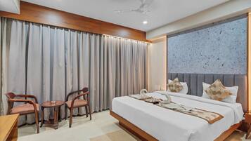 Quarto de hotel moderno com uma cama grande, duas cadeiras de madeira, uma mesinha redonda e um ventilador de teto.