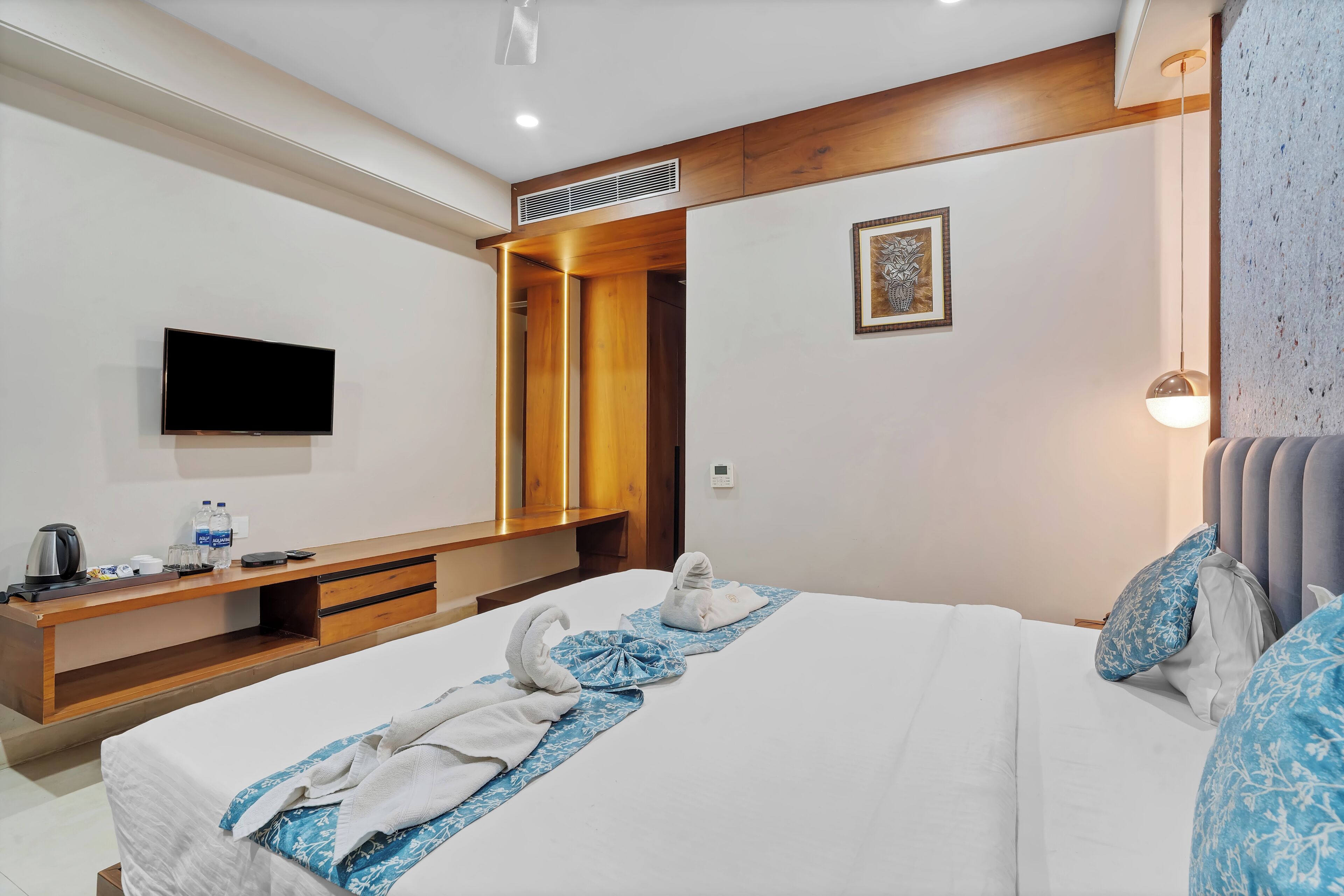 A hotel room with a bed, a flat-screen TV, a wooden console table, a wall-mounted TV, a framed picture, and a bedside lamp.