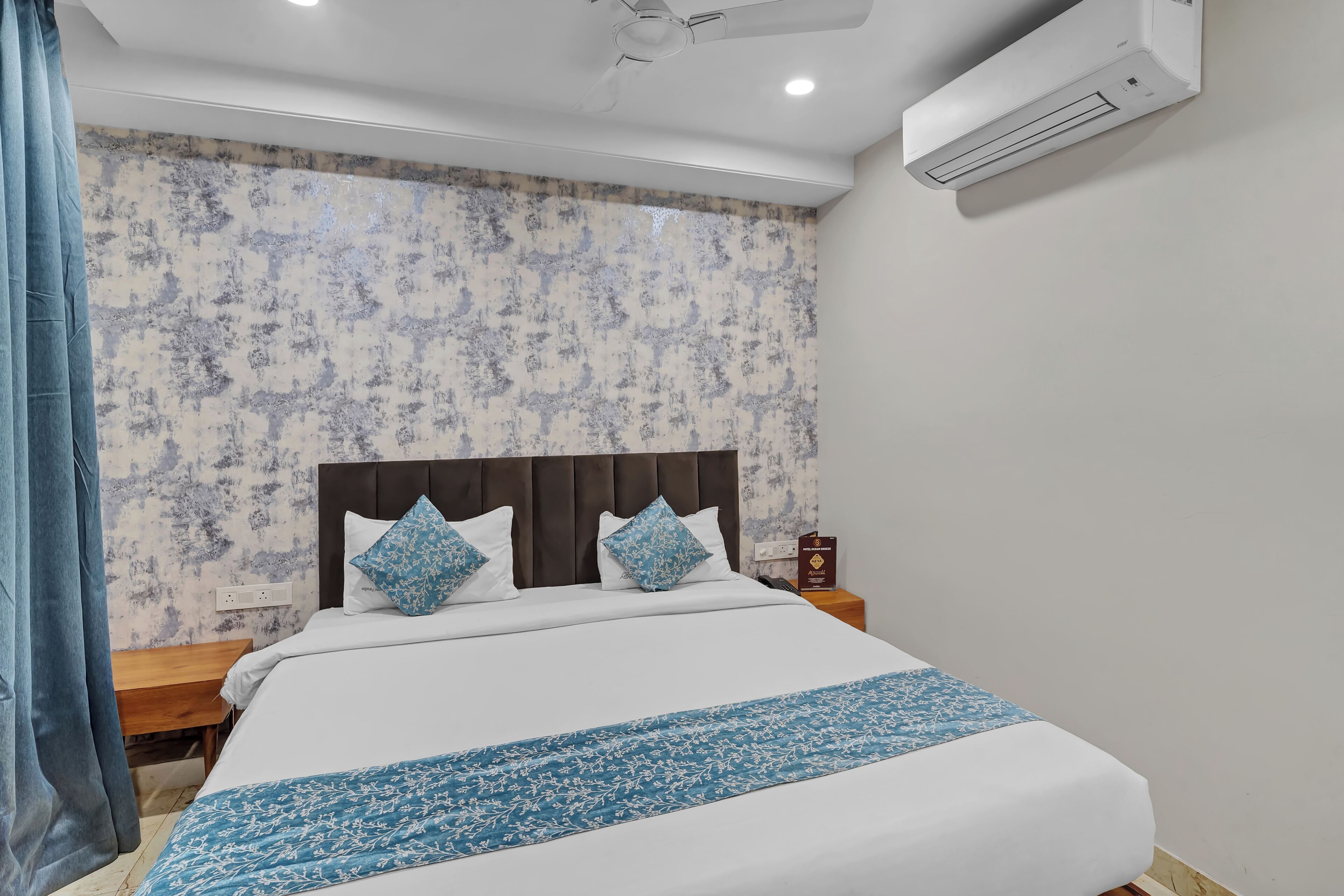 A neatly made bed with white and blue bedding, a dark headboard, and a patterned wall behind it.