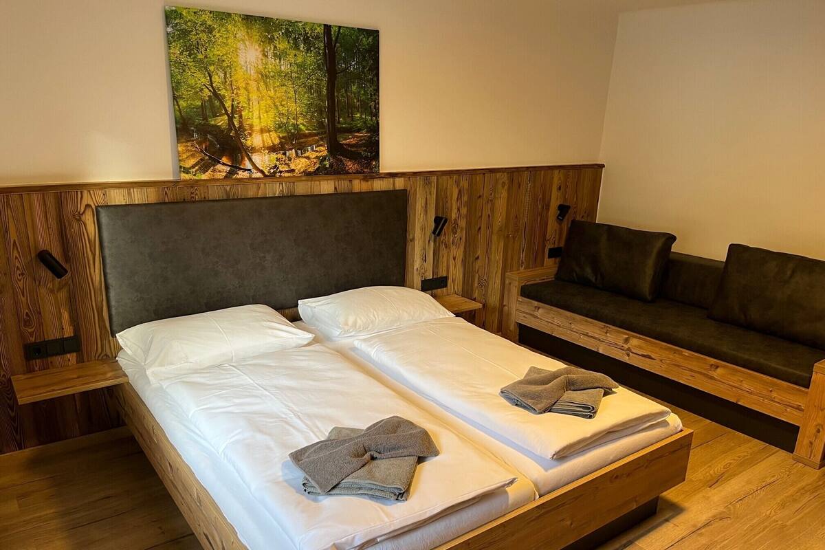 A double bed with white linens and two towels, a wooden headboard, a wooden bench with cushions, and a forest-themed painting on the wall.