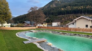 A swimming pool with a wooden deck, surrounded by a wooden fence, set against a backdrop of mountains and buildings. - Eibinghof Bed & Breakfast (Saalbach-Hinterglemm)
