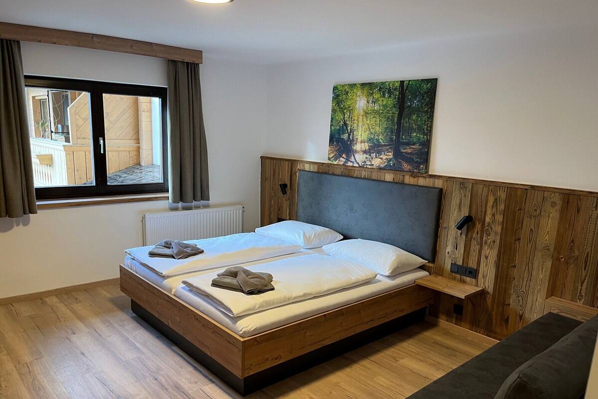 A double bed with a wooden headboard, a window with curtains, a radiator, and a painting of a forest scene.