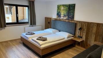 A double bed with a wooden headboard, a window with curtains, a radiator, and a painting of a forest scene.