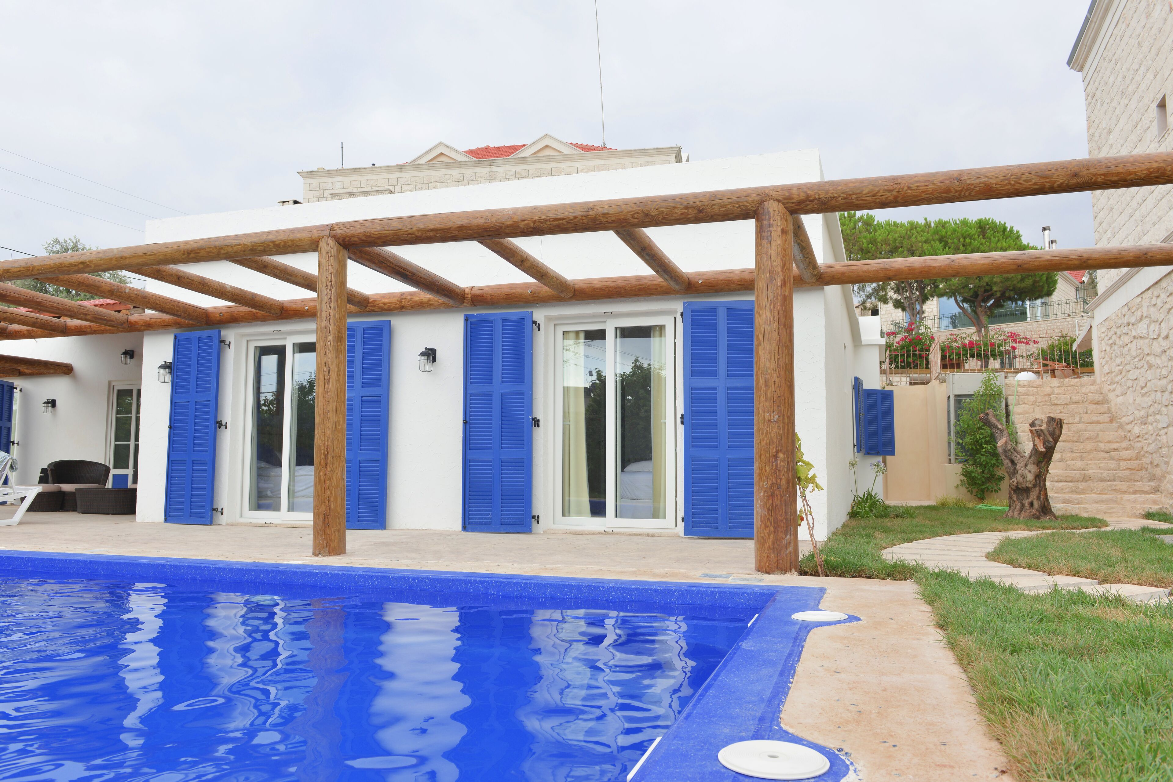 A poolside area with a pergola, a pool, and a building with blue shutters.