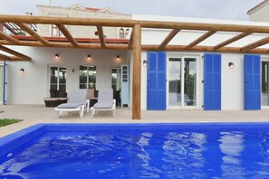 A poolside area with a wooden pergola, a pool, and a building with blue shutters and a white facade.