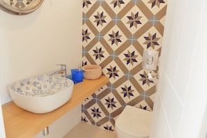 A bathroom with a white sink, a wooden shelf, a patterned tile wall, and a toilet.