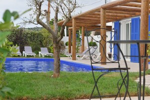Un área de piscina con sillones de descanso, una pérgola y un edificio con contraventanas azules.