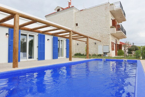 A swimming pool with a wooden pergola, a stone building with blue shutters, and a balcony with plants.