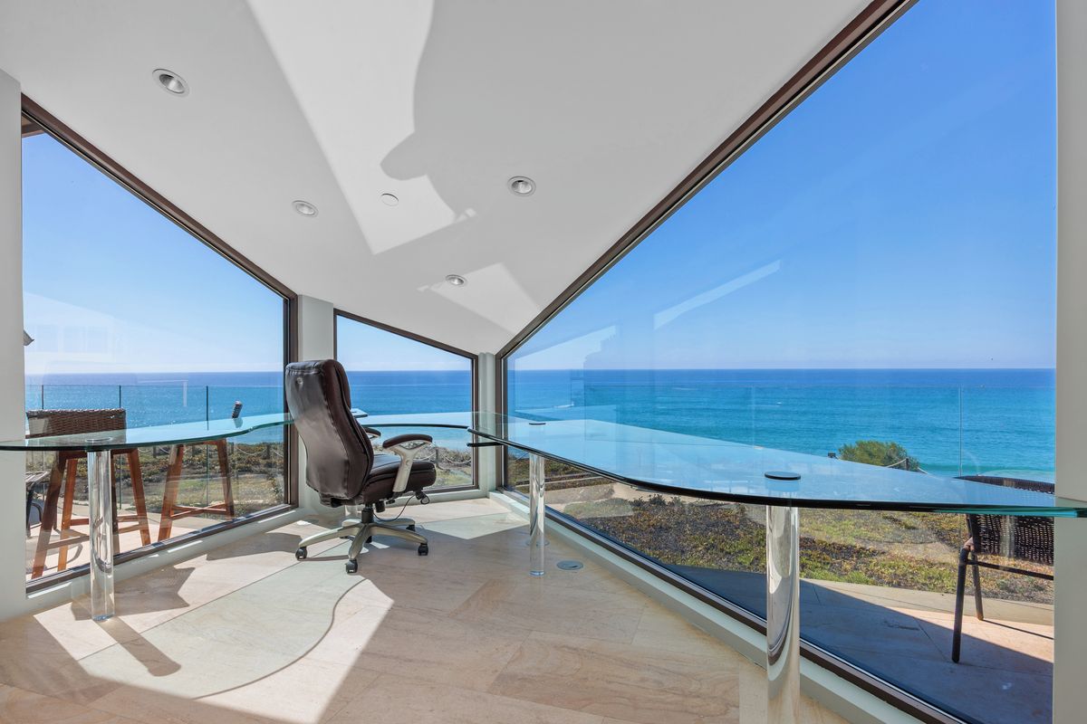 A modern office space with a panoramic ocean view, a glass table, and a single office chair.