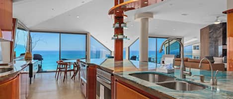 Una cocina moderna con una isla grande, un comedor con vistas al mar y un espacio de sala de estar con un sofá y una mesa de centro.