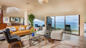 A spacious living room with a large window overlooking the ocean, a modern chandelier, and a cozy seating area with a sofa and chairs. - Enchanting Oasis Stunning Luxury Oceanfront Condo -180° Ocean Views + Pool (Solana Beach)