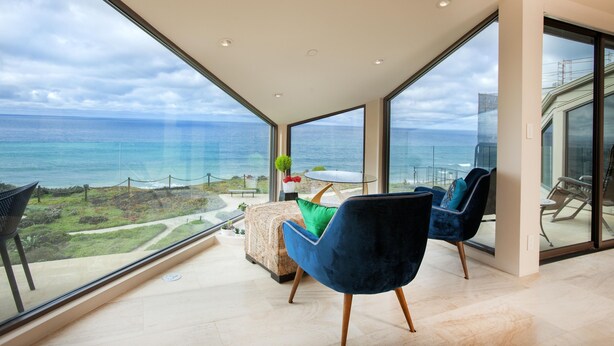 A modern living room with large windows overlooking the ocean, a cozy seating area, and a view of the sea. - Enchanting Oasis Stunning Luxury Oceanfront Condo -180° Ocean Views + Pool (Solana Beach)