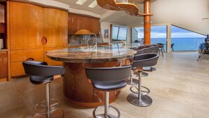 A modern kitchen with a central island, bar stools, and a view of the ocean. - Enchanting Oasis Stunning Luxury Oceanfront Condo -180° Ocean Views + Pool (Solana Beach)