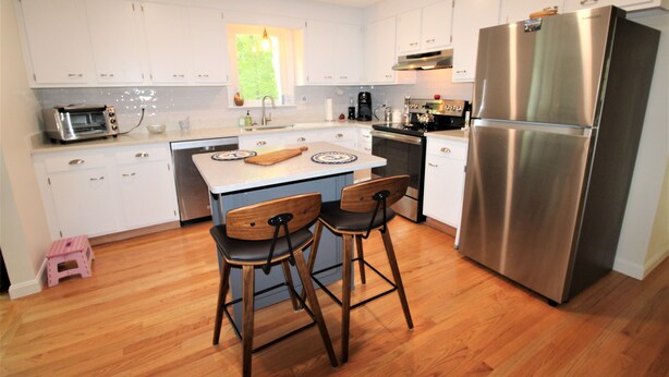 A modern kitchen with white cabinets, a stainless steel refrigerator, a built-in oven, and a coffee maker. There are two wooden chairs at the island and a small pink stool. - Great Home in a Quiet Neighborhood Yet Close to Town and Beaches (2312) (Dennis)