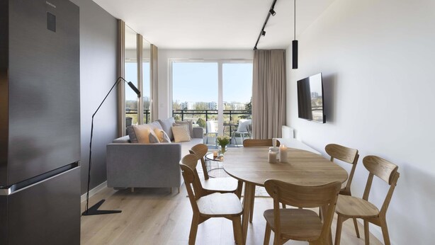 A modern living room with a grey sofa, wooden dining table, and chairs. A large window offers a view of the city. - Elite Apartments Seastory (Gdansk)