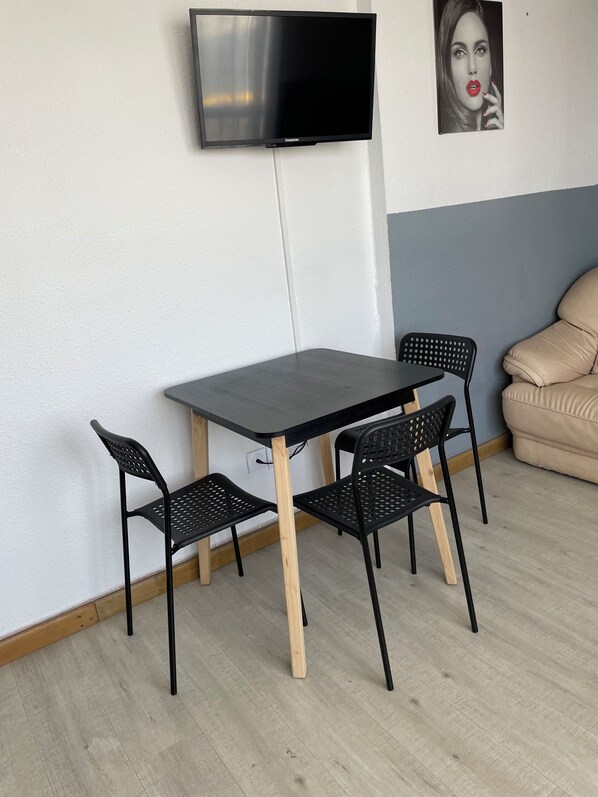En hyggelig spiseplads med et bord og fire stole, et fladskærms-tv på væggen og en beige sofa i baggrunden.