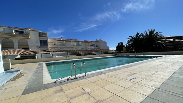 Una piscina con borde de baldosas, rodeada de edificios con balcones y un cielo azul despejado.