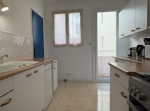 A kitchen with white cabinets, a blue door, and a window. - Appartement T3 vue mer (Toulon)