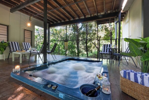 A hot tub with a wooden deck, surrounded by lounge chairs and a table, with a view of trees through large windows.