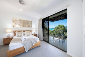 A bedroom with a large bed, bedside tables, a window with blinds, and a balcony with outdoor furniture.